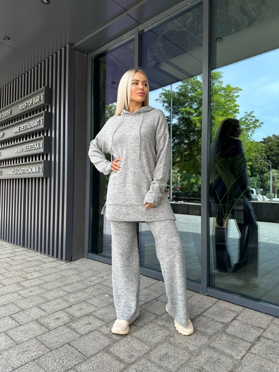Woman in a gray tracksuit standing outside a modern building with glass doors.
