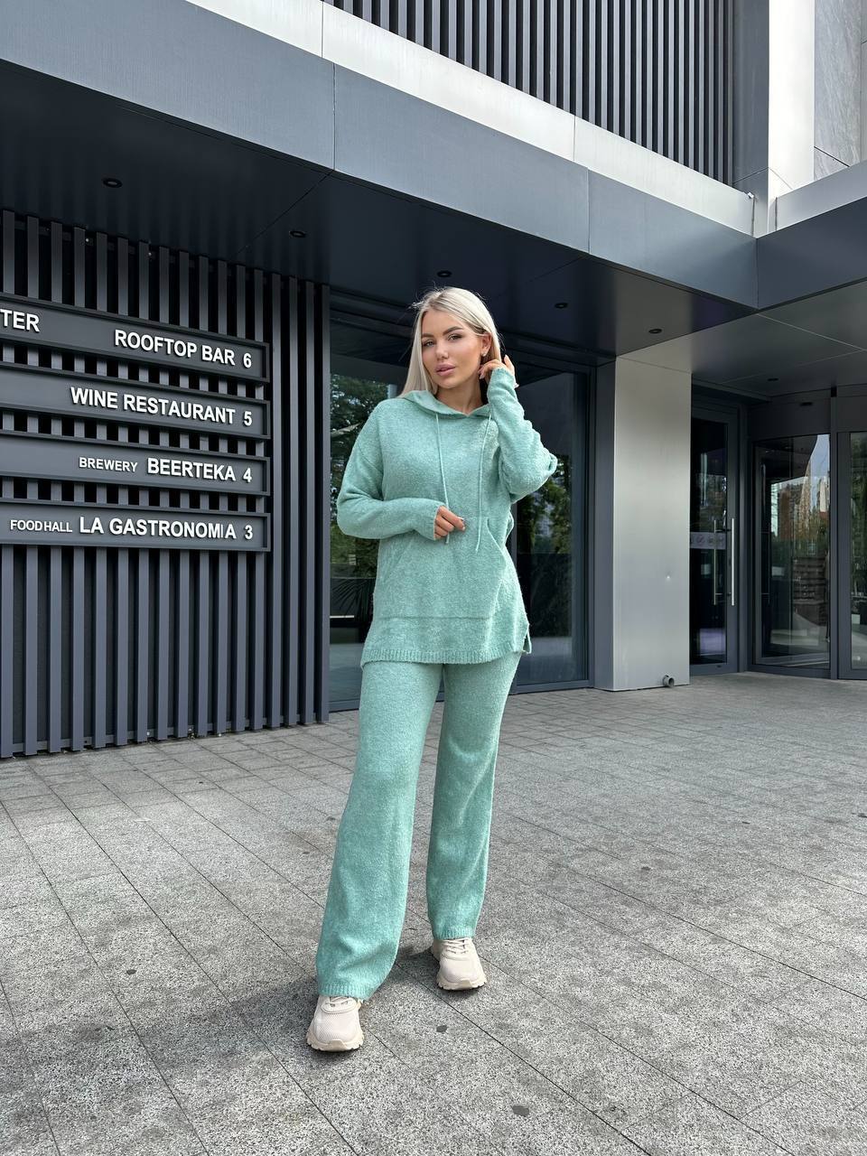 Woman in a teal tracksuit standing in front of a modern building with signage.