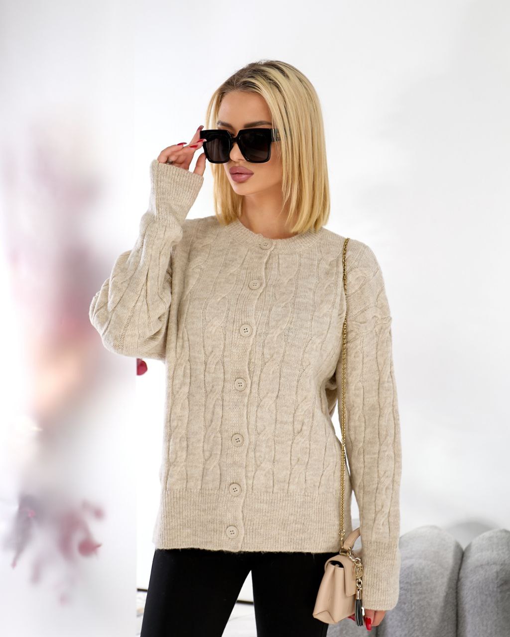 Woman wearing a beige cable knit sweater with sunglasses and a handbag on a blurred background