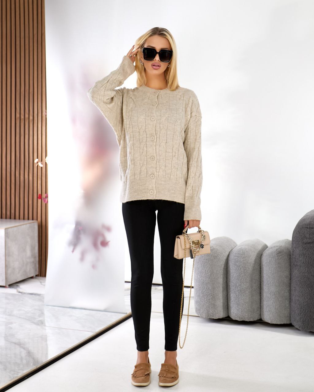 Woman wearing a beige sweater, black leggings, and brown shoes in a modern interior setting.