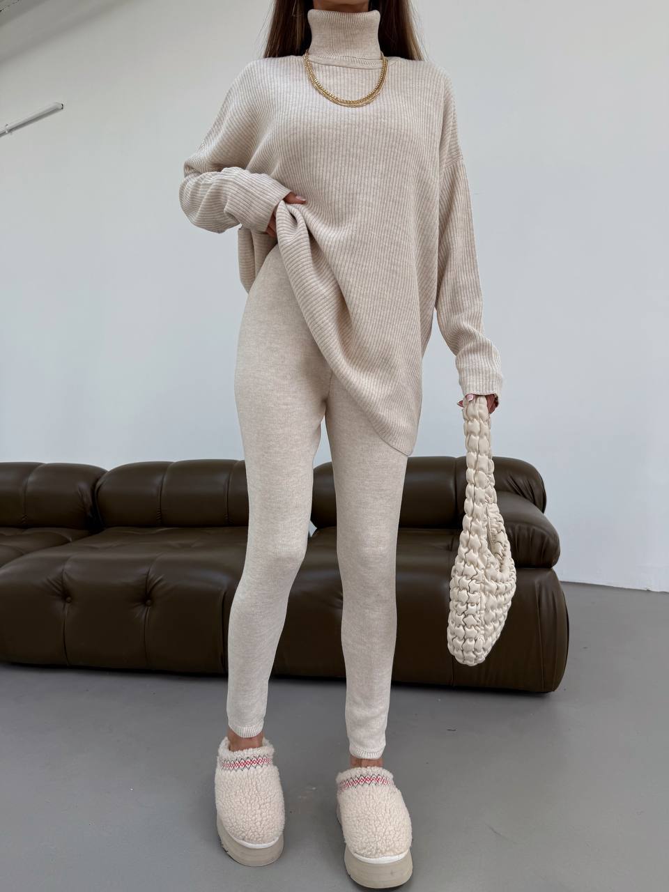 Person wearing a beige knit outfit with white shoes, holding a matching bag in a minimalistic room.