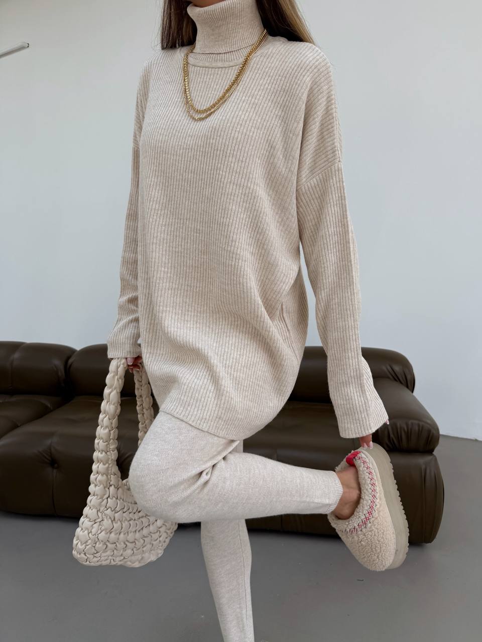 Person wearing a beige turtleneck sweater and pants, holding a woven handbag, with a brown sofa in the background.