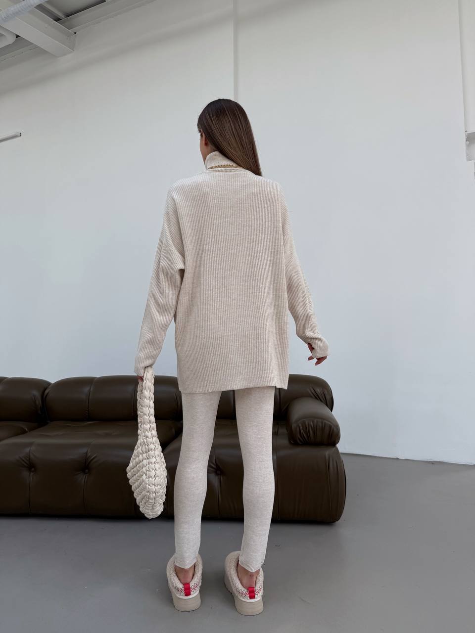 Person wearing a beige sweater and leggings standing in a minimalistic room with a brown sofa.