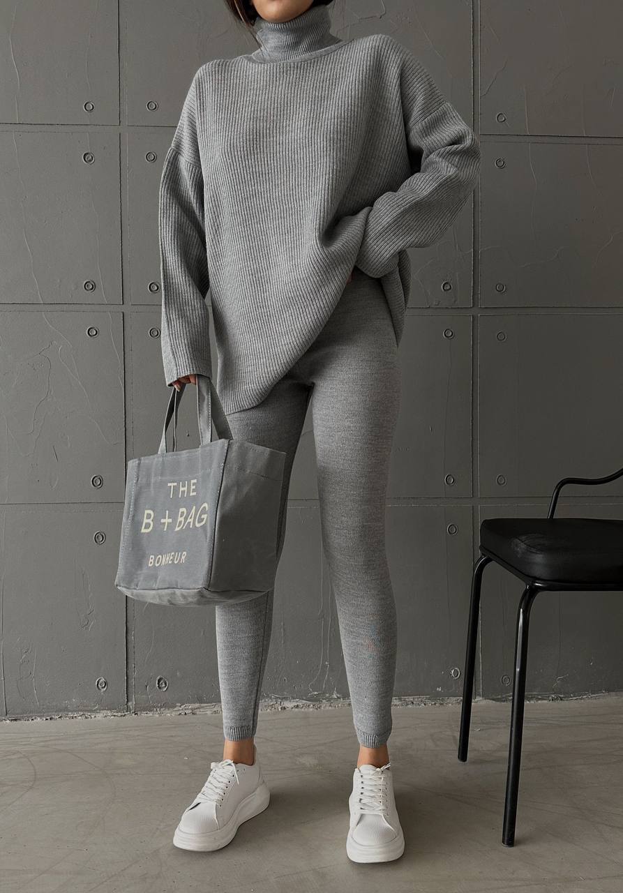 Person wearing a gray outfit holding a bag with text against a gray wall.
