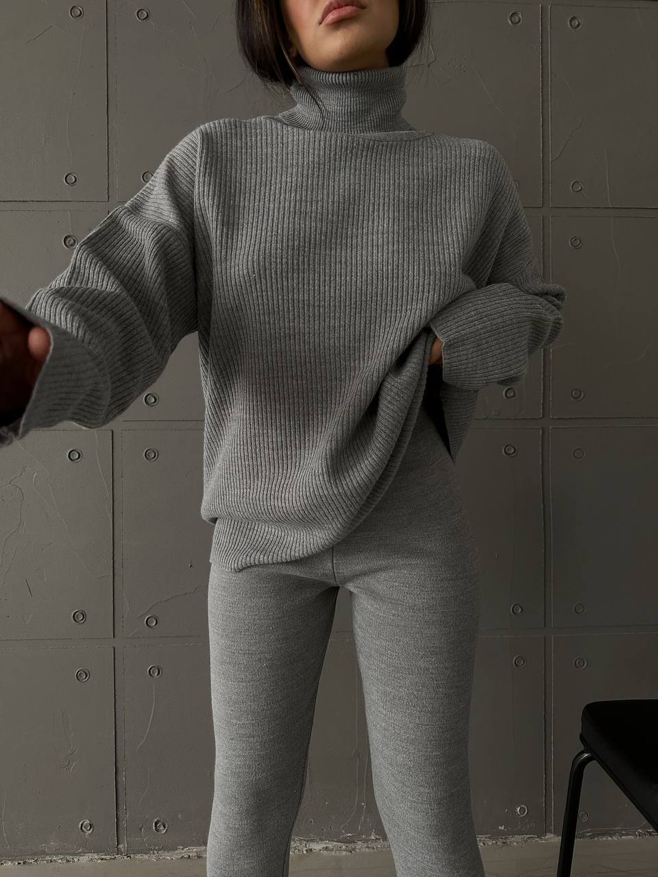 Person wearing a gray ribbed sweater and pants against a textured wall.
