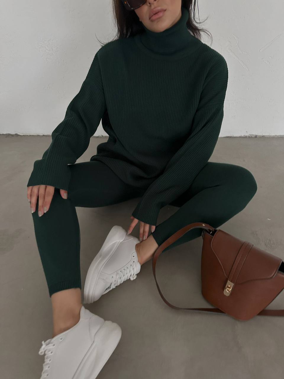 Person wearing a dark green turtleneck and leggings with white sneakers, sitting on the floor with a brown handbag.