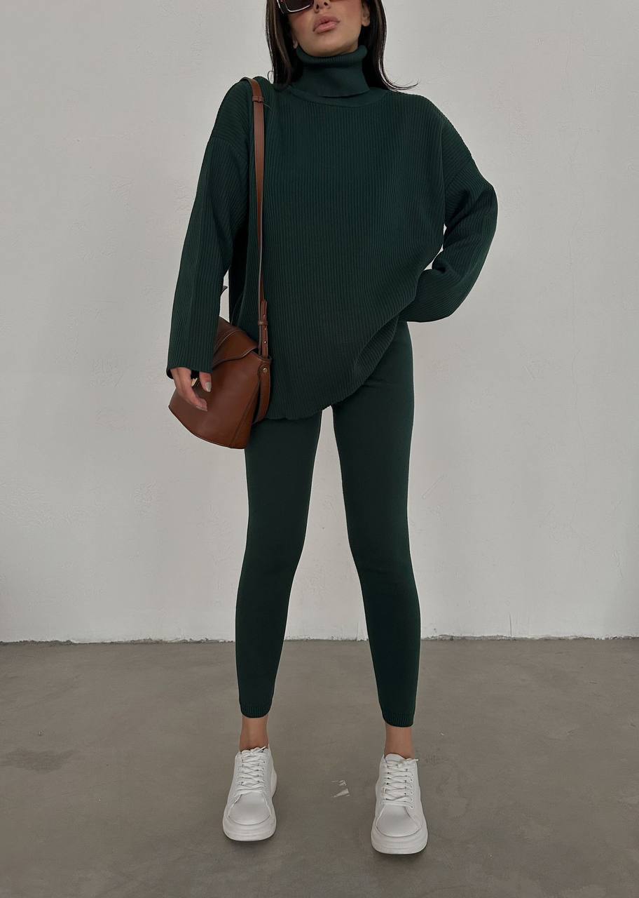 Person wearing a dark green outfit with white sneakers and a brown bag against a plain background