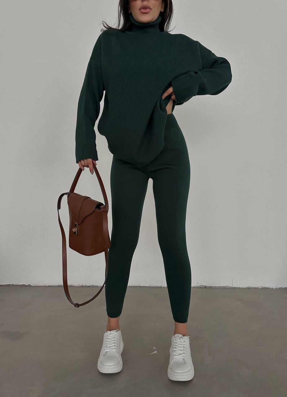 Person wearing a dark green outfit with a brown bag against a plain background