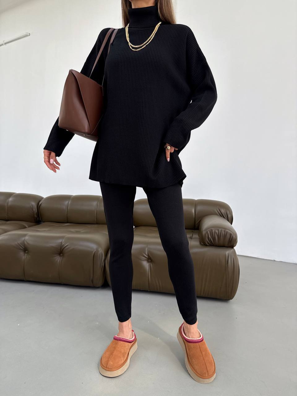 Person wearing a black outfit with brown shoes and a brown bag in a minimalistic room.