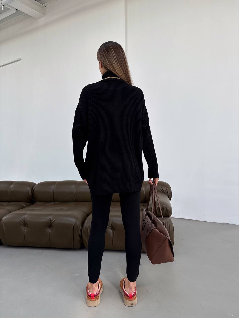 Person in a black outfit holding a brown bag in a minimalistic room with a brown sofa.