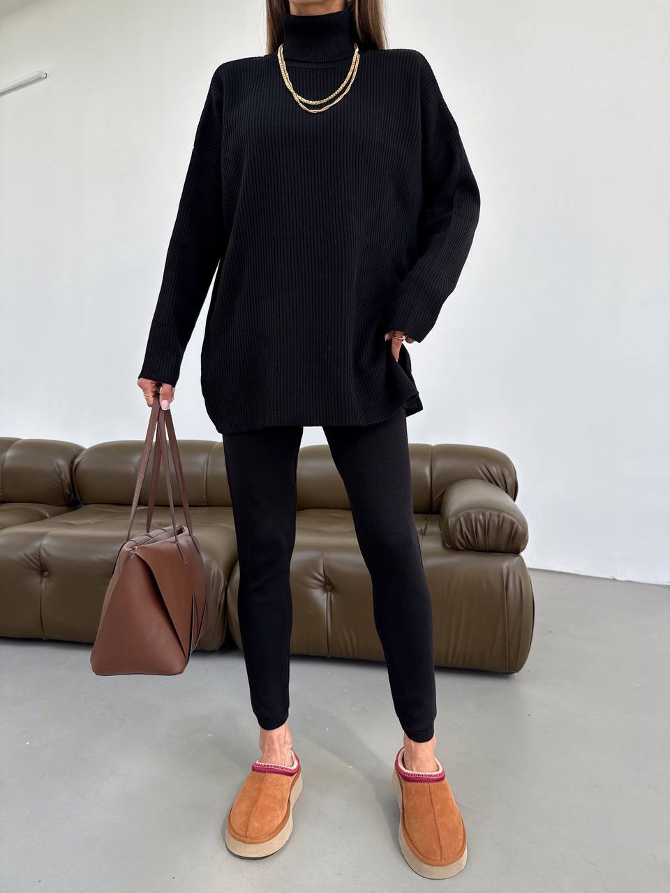Person wearing a black outfit with brown shoes and a brown bag, standing in front of a brown sofa.