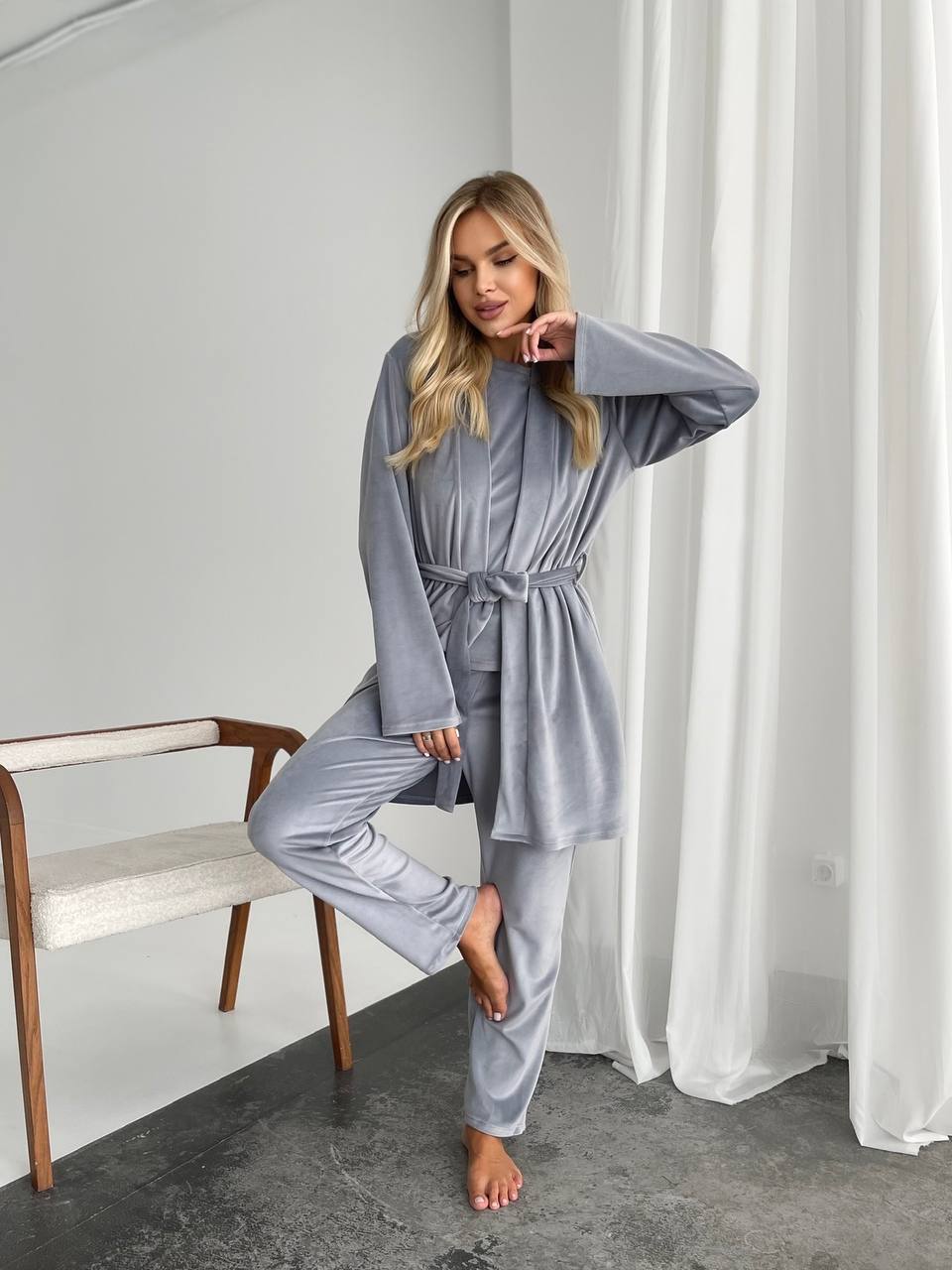 Woman in gray pajama set standing in a room with white curtains and a wooden chair.