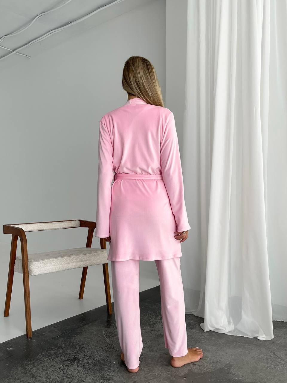 Person wearing a pink pajama set standing in a room with a chair and white curtains.
