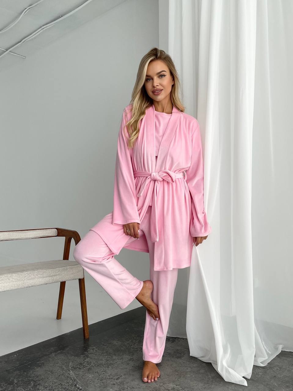 Woman wearing a pink robe and pants in a room with a bench and white curtains.