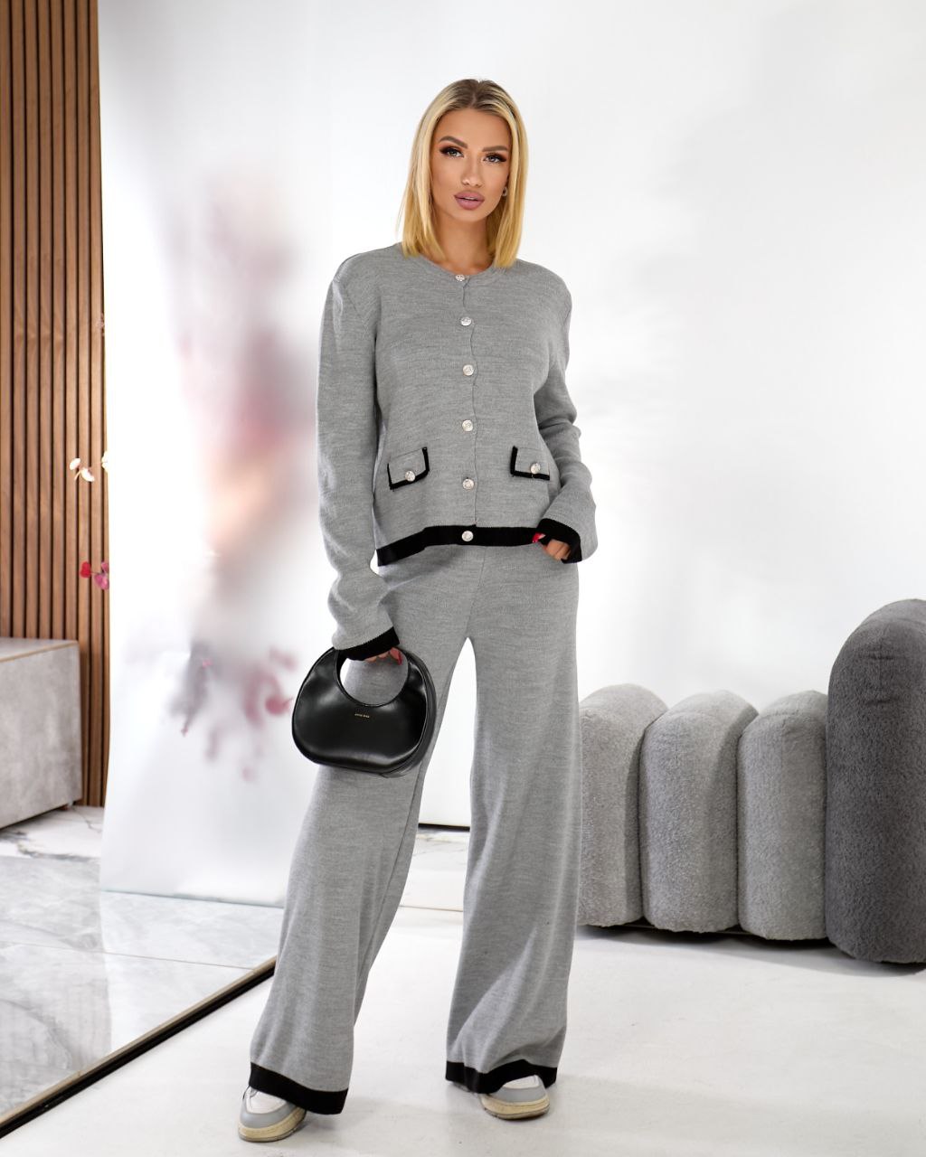 Woman in a gray outfit holding a black handbag in a modern living room.