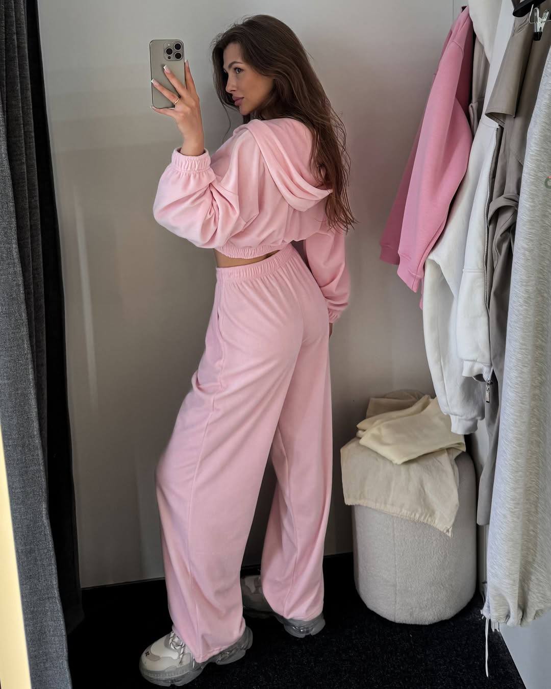 Woman in a pink tracksuit taking a mirror selfie in a room with clothes on a rack.