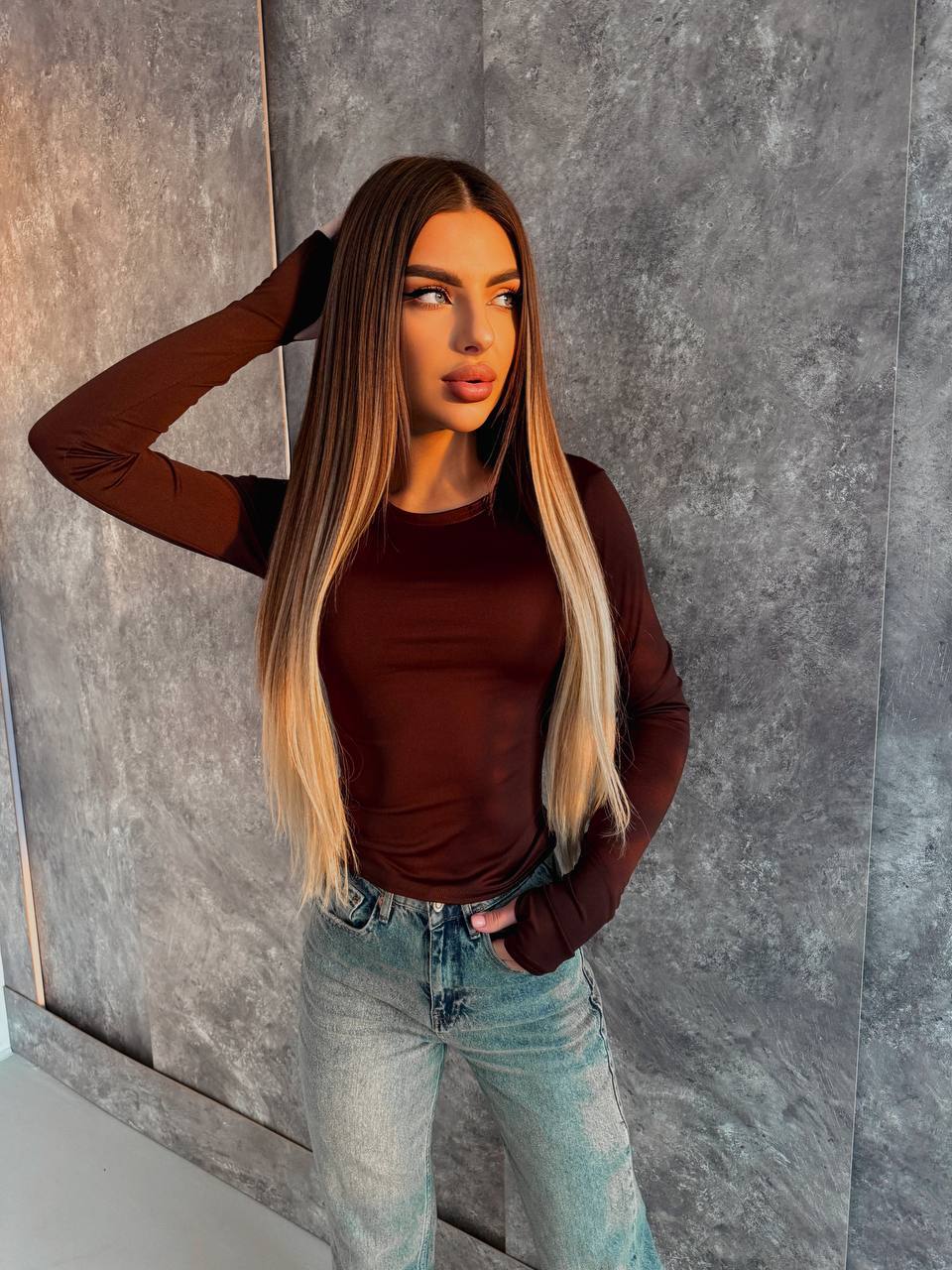 Person wearing a brown long-sleeve top and light blue jeans against a gray textured wall.