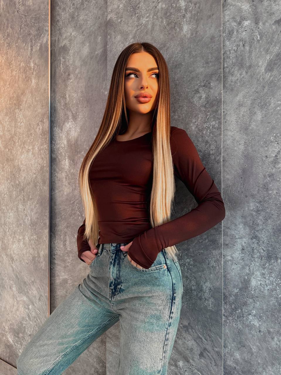 Woman wearing a brown top and blue jeans against a gray wall