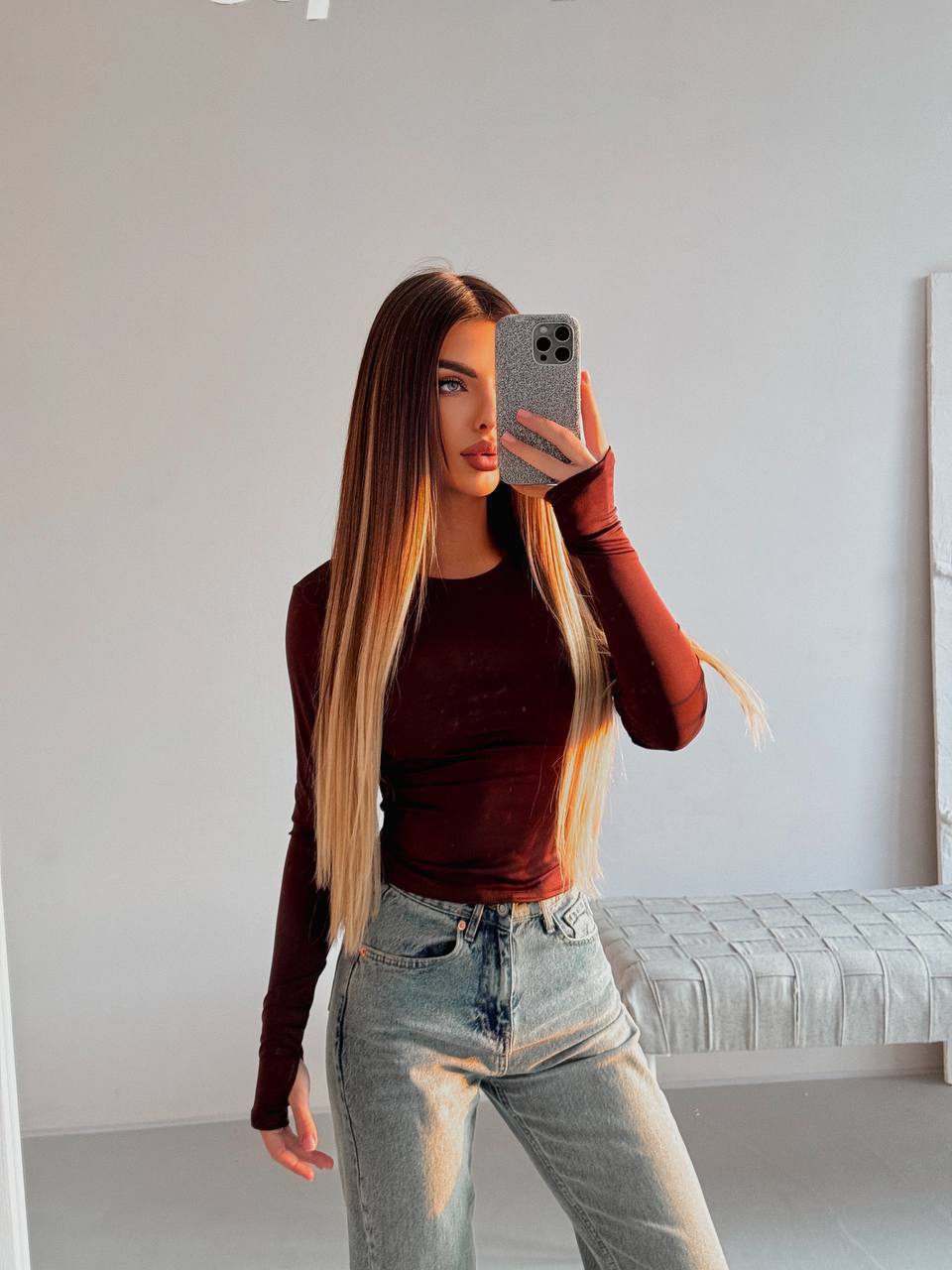 Woman taking a mirror selfie wearing a brown long-sleeve top and light blue jeans.