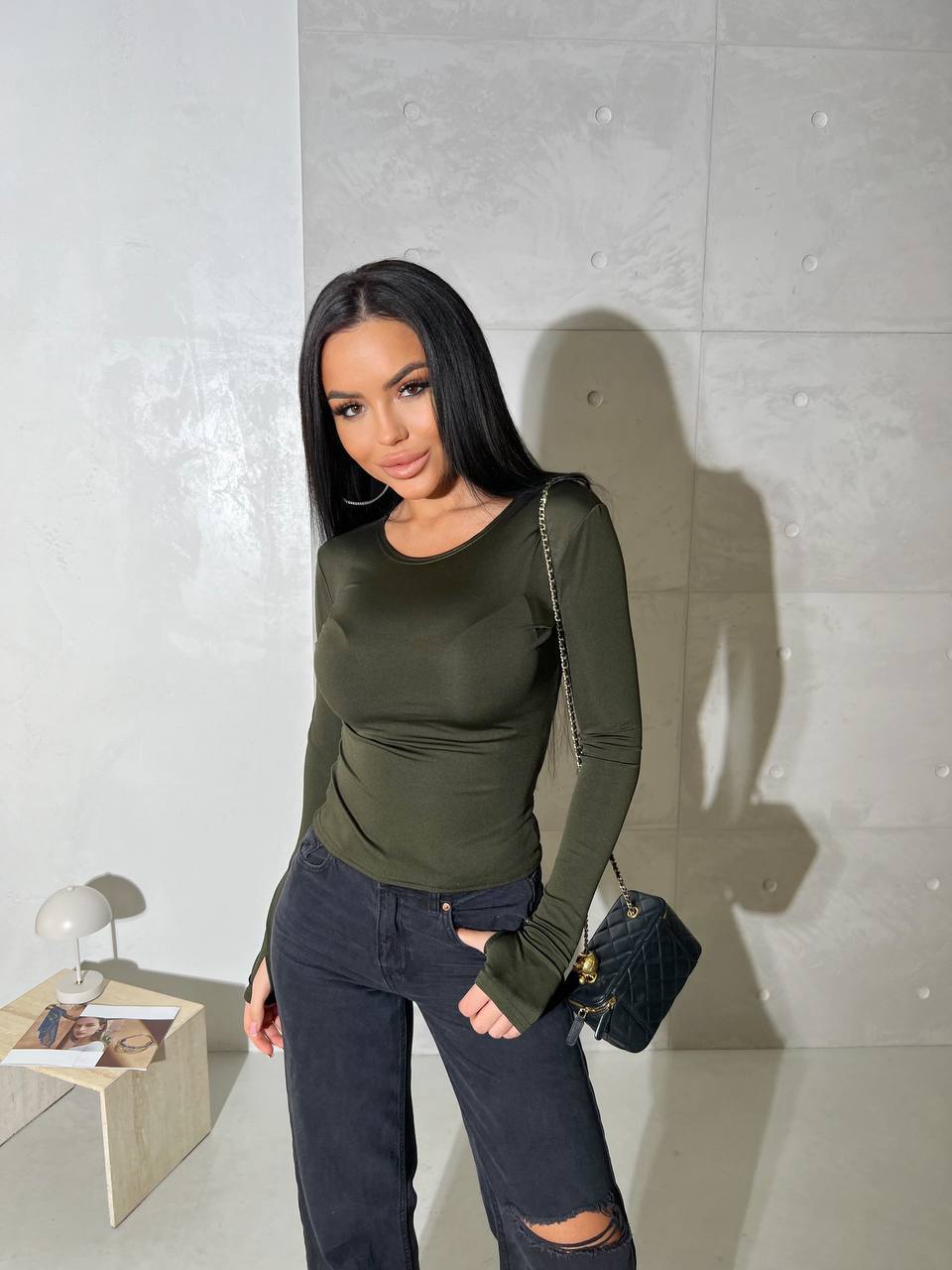 Woman wearing an olive green long-sleeve top and dark jeans against a light gray wall.