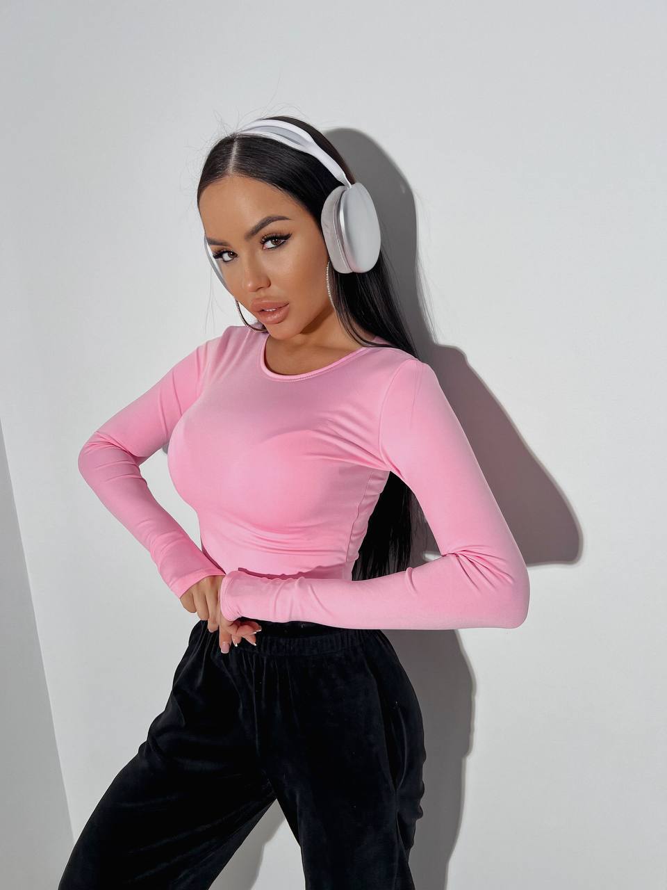 Person wearing a pink long-sleeve top and black pants with white headphones on a plain background