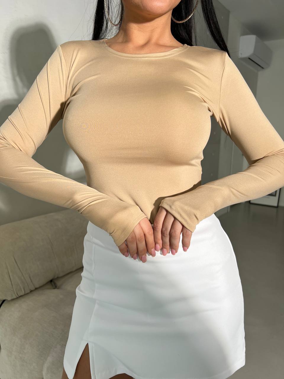 Person wearing a beige long-sleeve top and white skirt indoors.