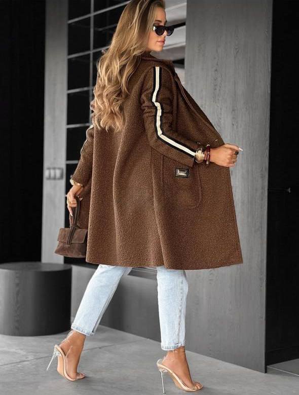 Woman wearing a brown coat with white stripes, light blue jeans, and high heels in an urban setting.
