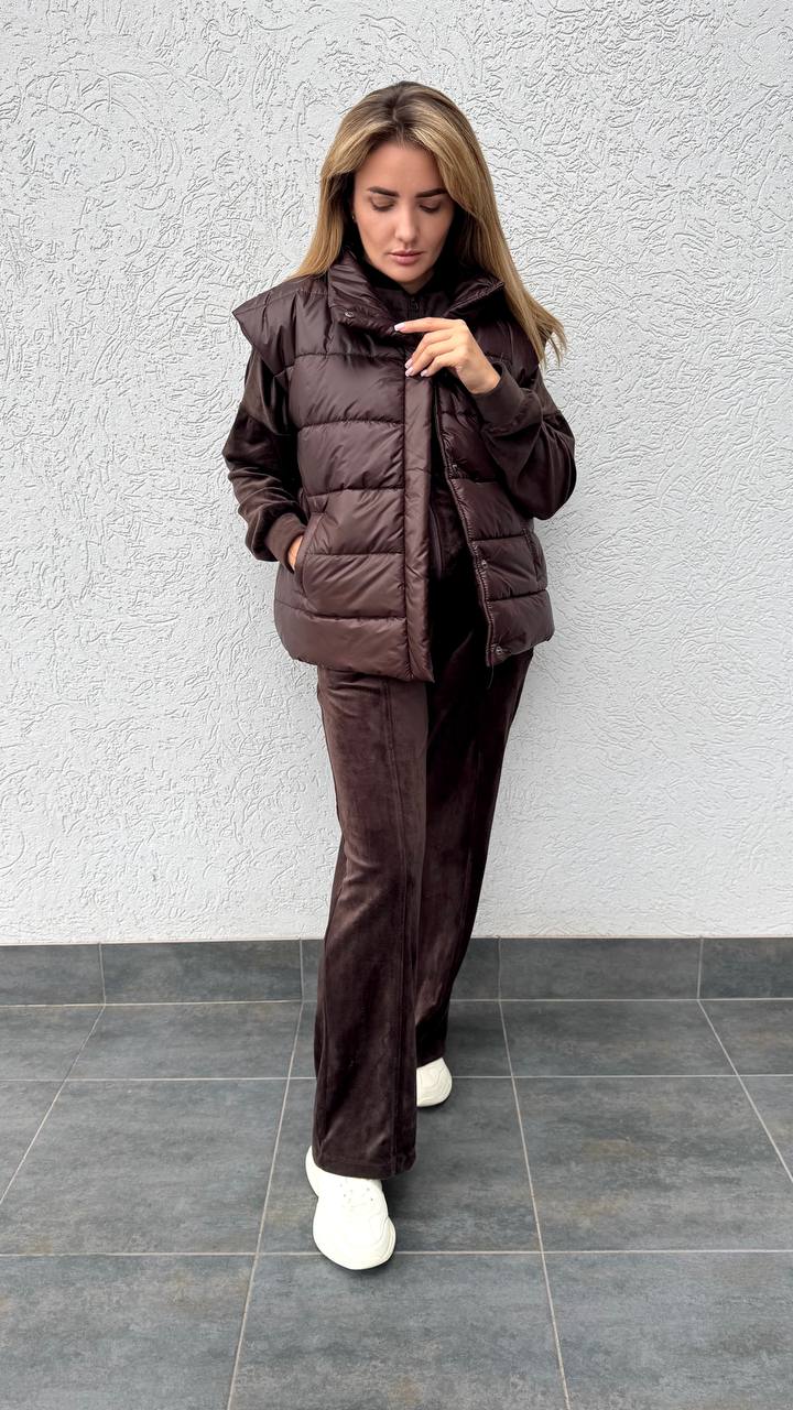 Woman wearing a brown puffer jacket and pants against a gray wall.