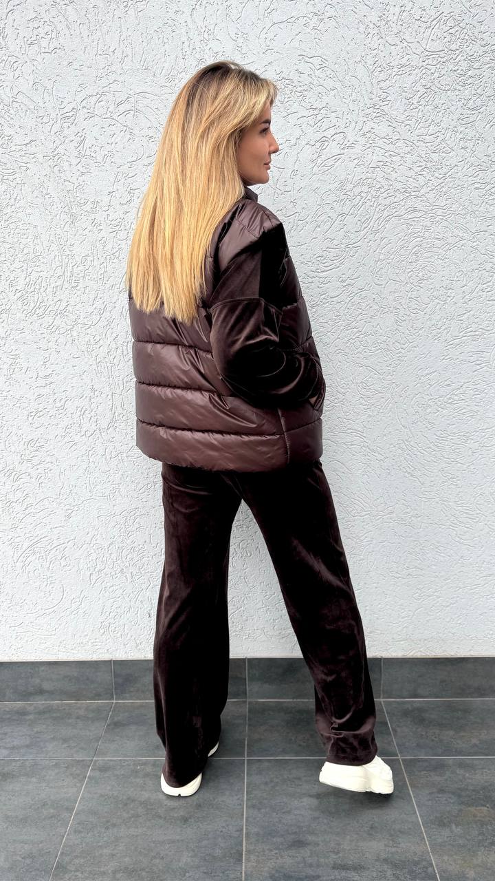 Woman wearing a brown puffer jacket and pants against a light gray wall.