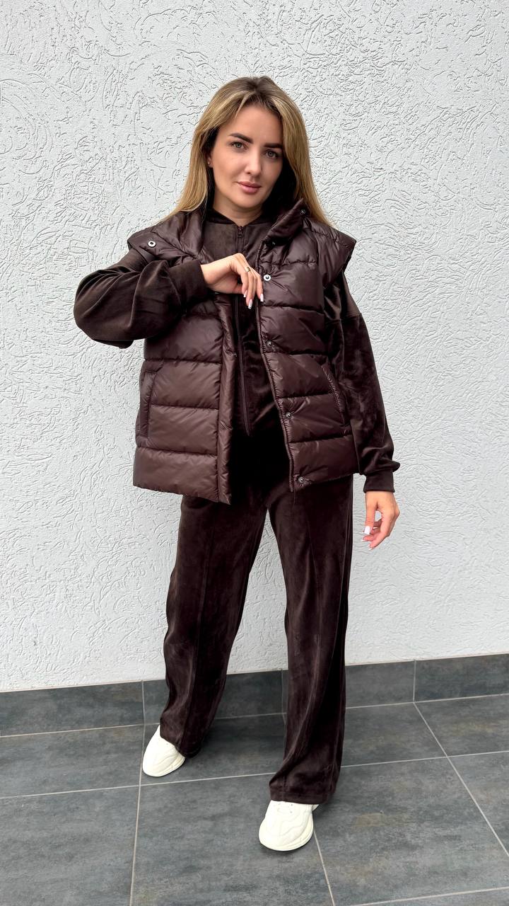 Woman wearing a brown puffer jacket and pants against a gray wall.