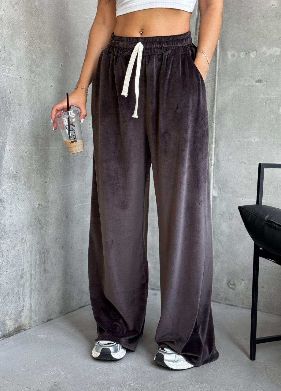 Person wearing brown wide-leg pants with a white top and silver shoes, holding a drink against a gray wall.