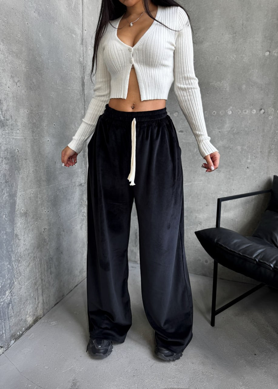 Person wearing a white ribbed cardigan and black pants against a gray wall.