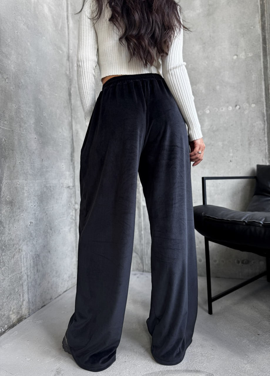 Person wearing black wide-leg pants and a light-colored sweater against a gray wall.
