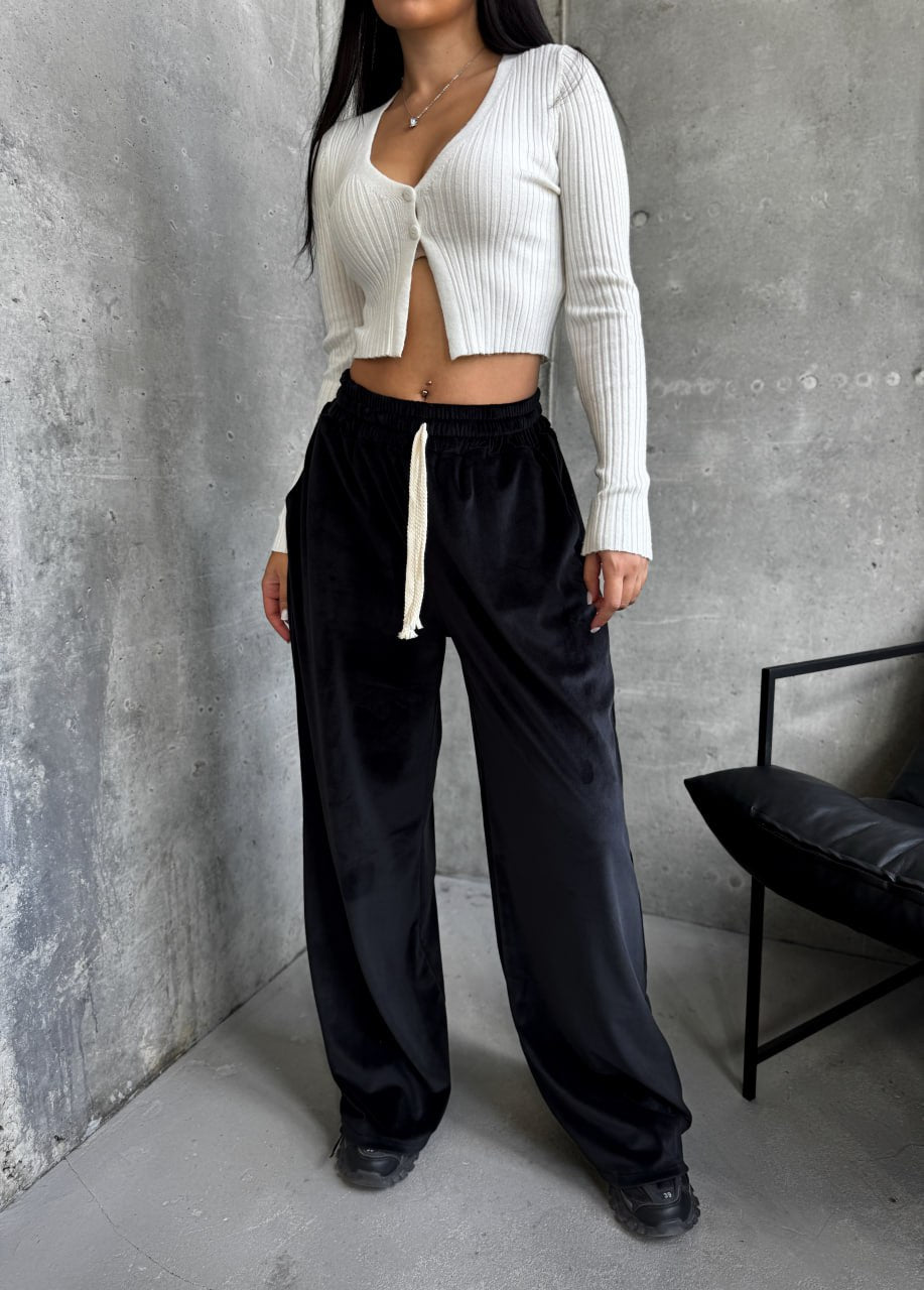 Person wearing a white cropped sweater and black pants against a gray concrete wall.