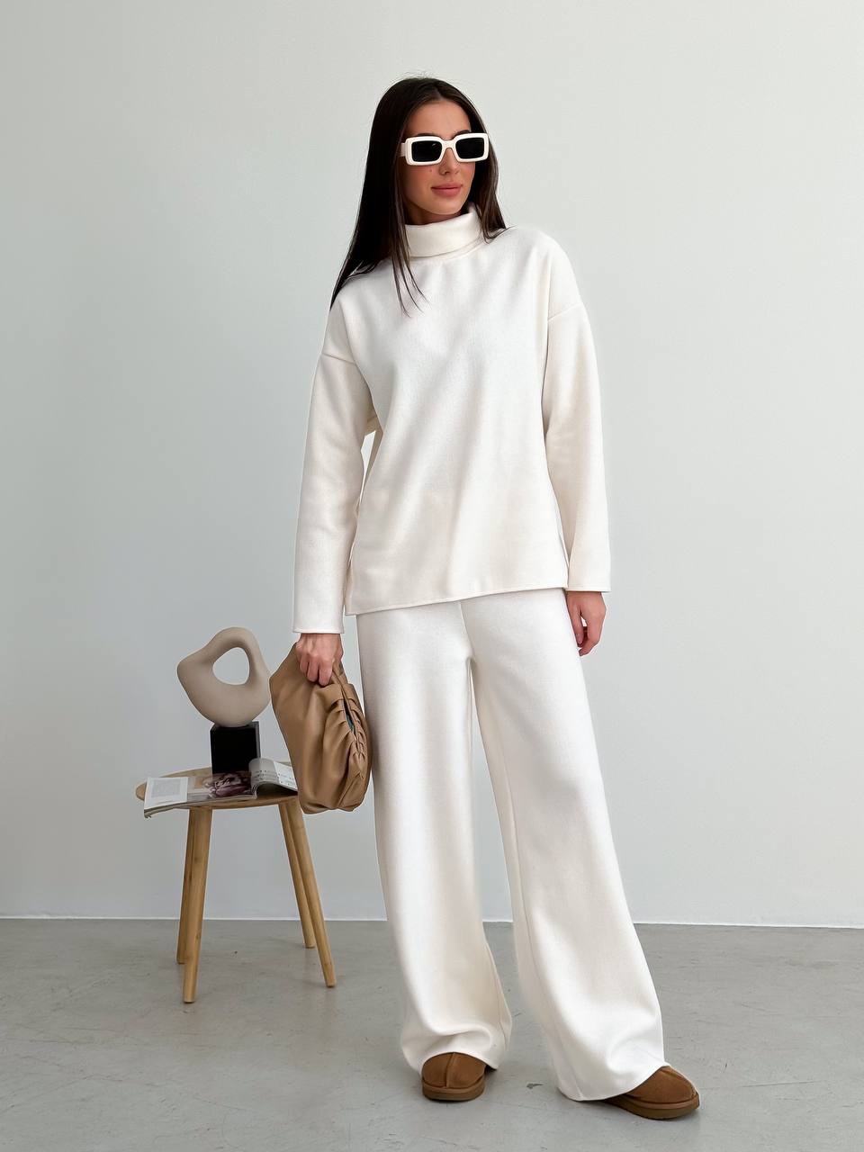 Woman in a white outfit standing in a minimalistic room with a chair and bag.