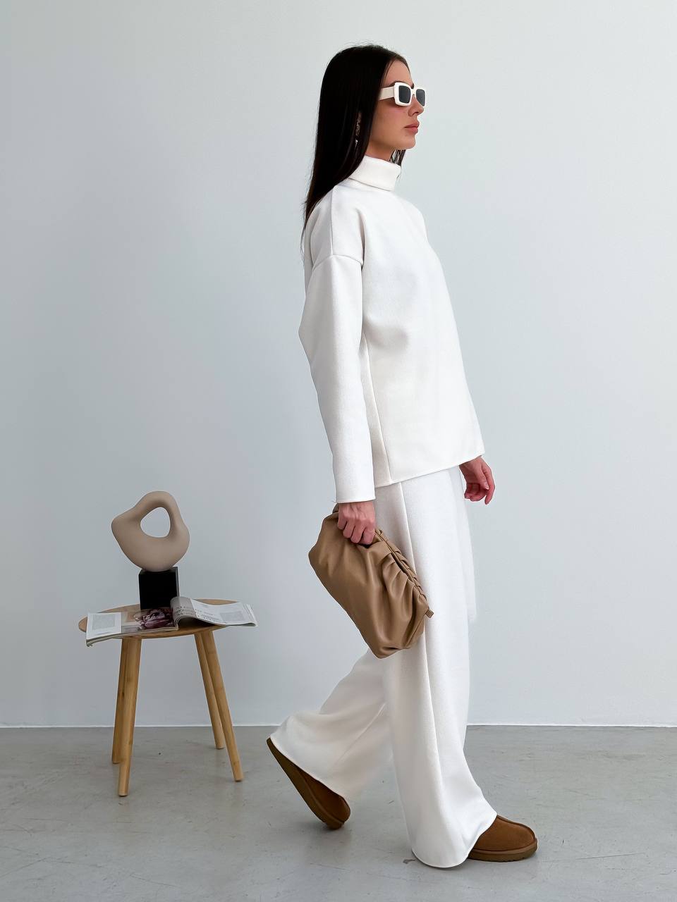 Person wearing a white outfit with a beige bag and shoes, standing in a minimalistic room.