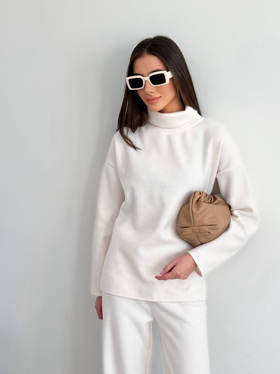 Woman wearing a white outfit with sunglasses and a beige clutch against a light gray background