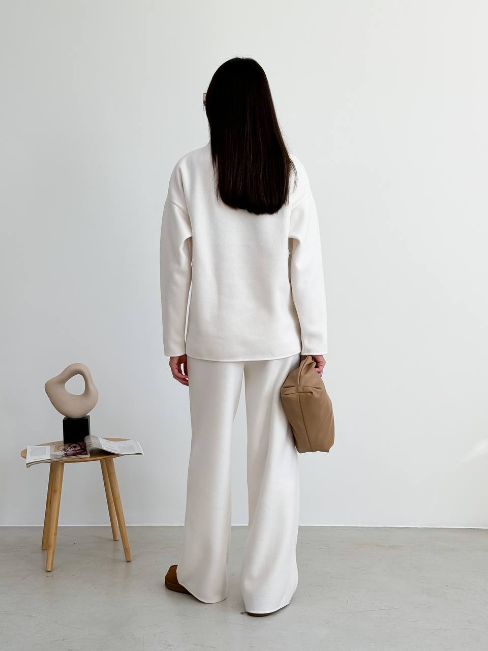 Person wearing a white outfit standing in a minimalistic room with a small table and chair.
