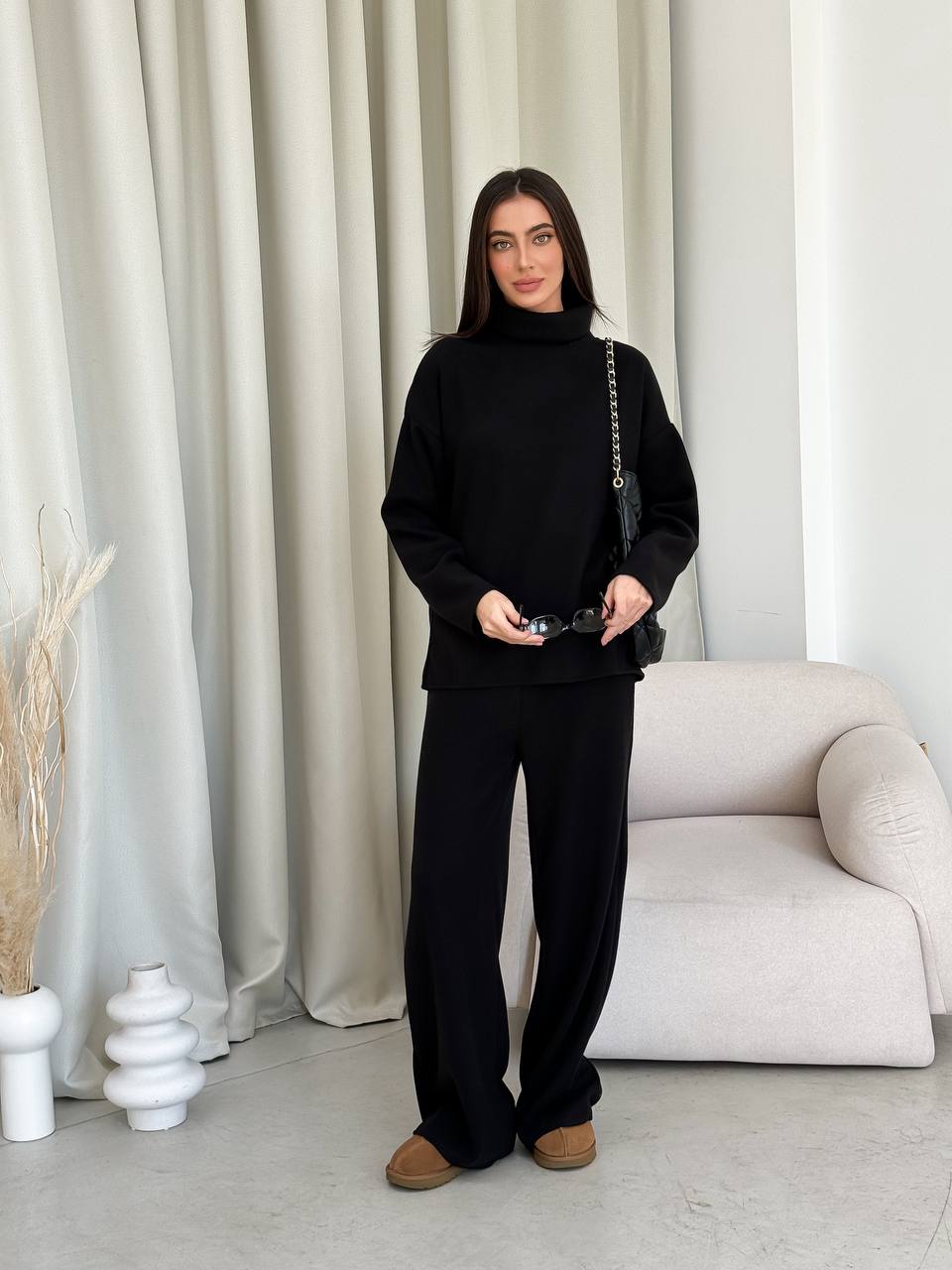 Person wearing a black outfit standing in a minimalistic room with white curtains and a white sofa.