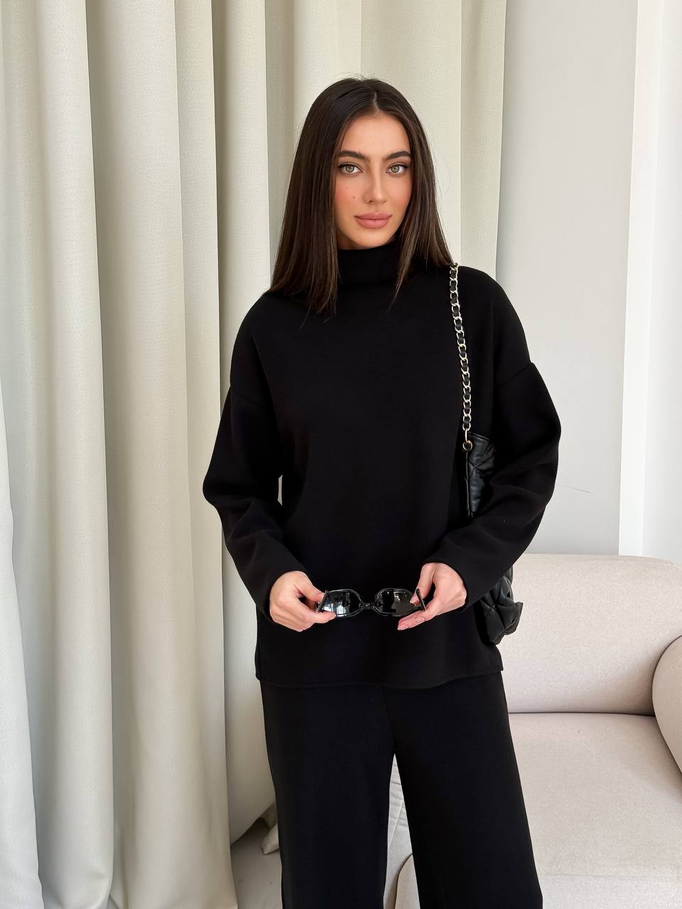 Woman in a black outfit holding sunglasses in front of a neutral curtain.