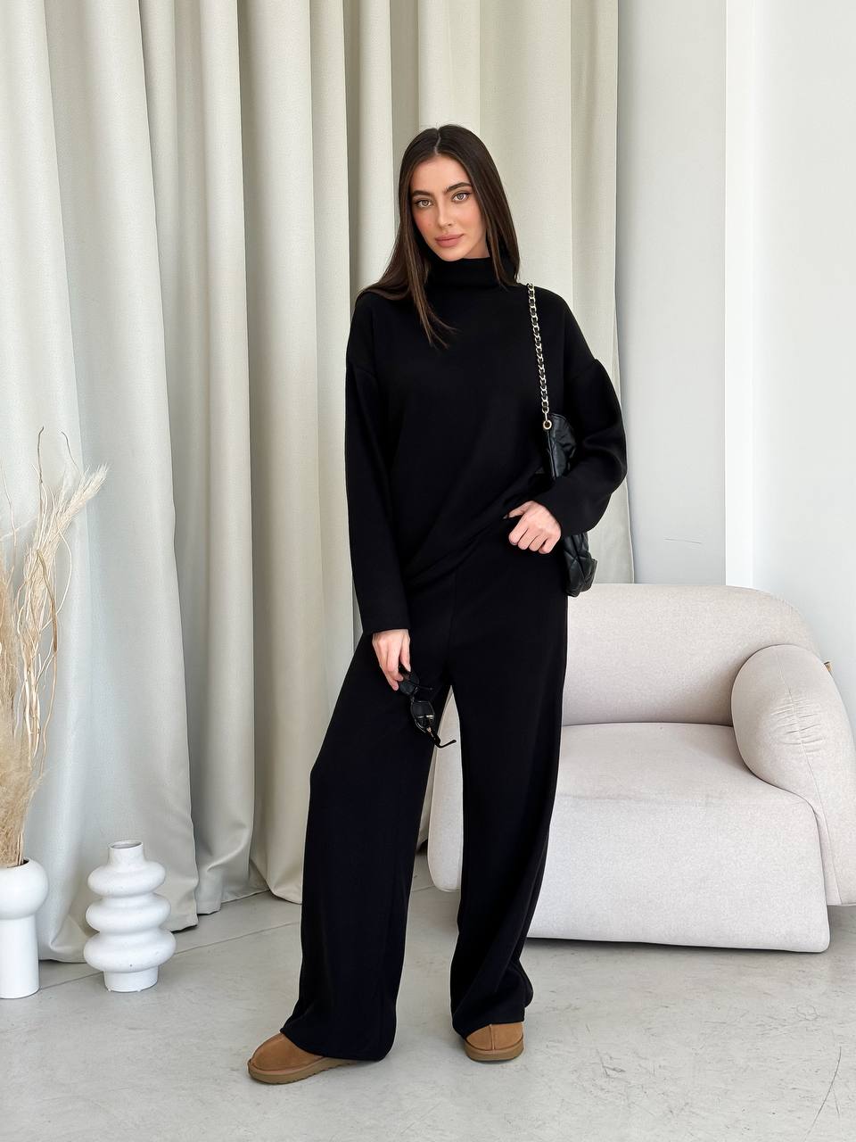 Woman in a black outfit standing in a minimalistic room with white curtains and a white sofa.