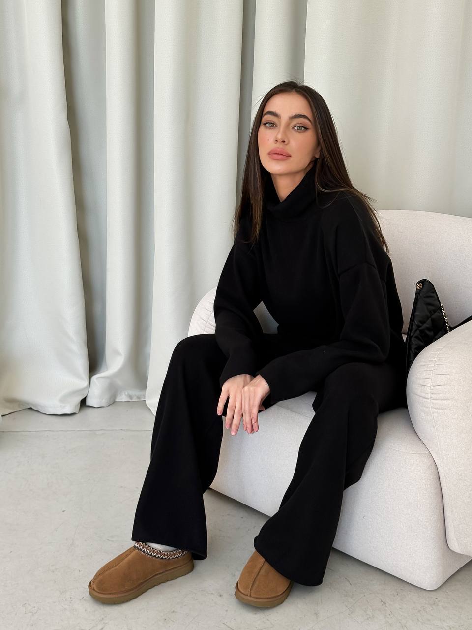 Woman in black outfit sitting on a white couch with white curtains in the background
