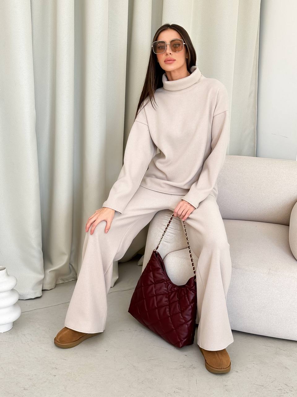 Woman in a beige outfit sitting on a couch with a maroon handbag.
