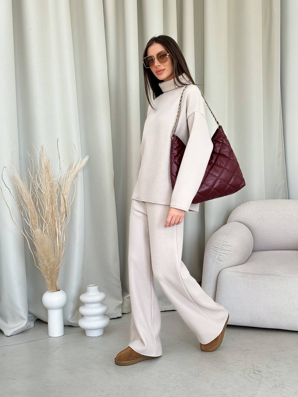 Woman in a stylish outfit with a maroon handbag in a room with curtains and decor.