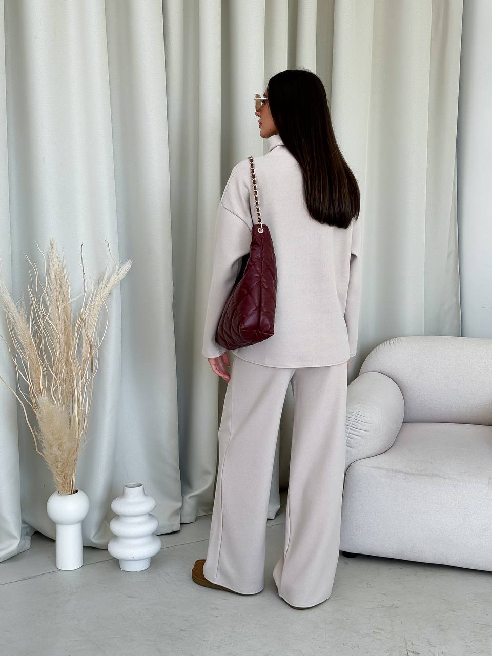 Woman in a light gray outfit holding a red bag in a minimalistic room with white curtains and a white sofa.