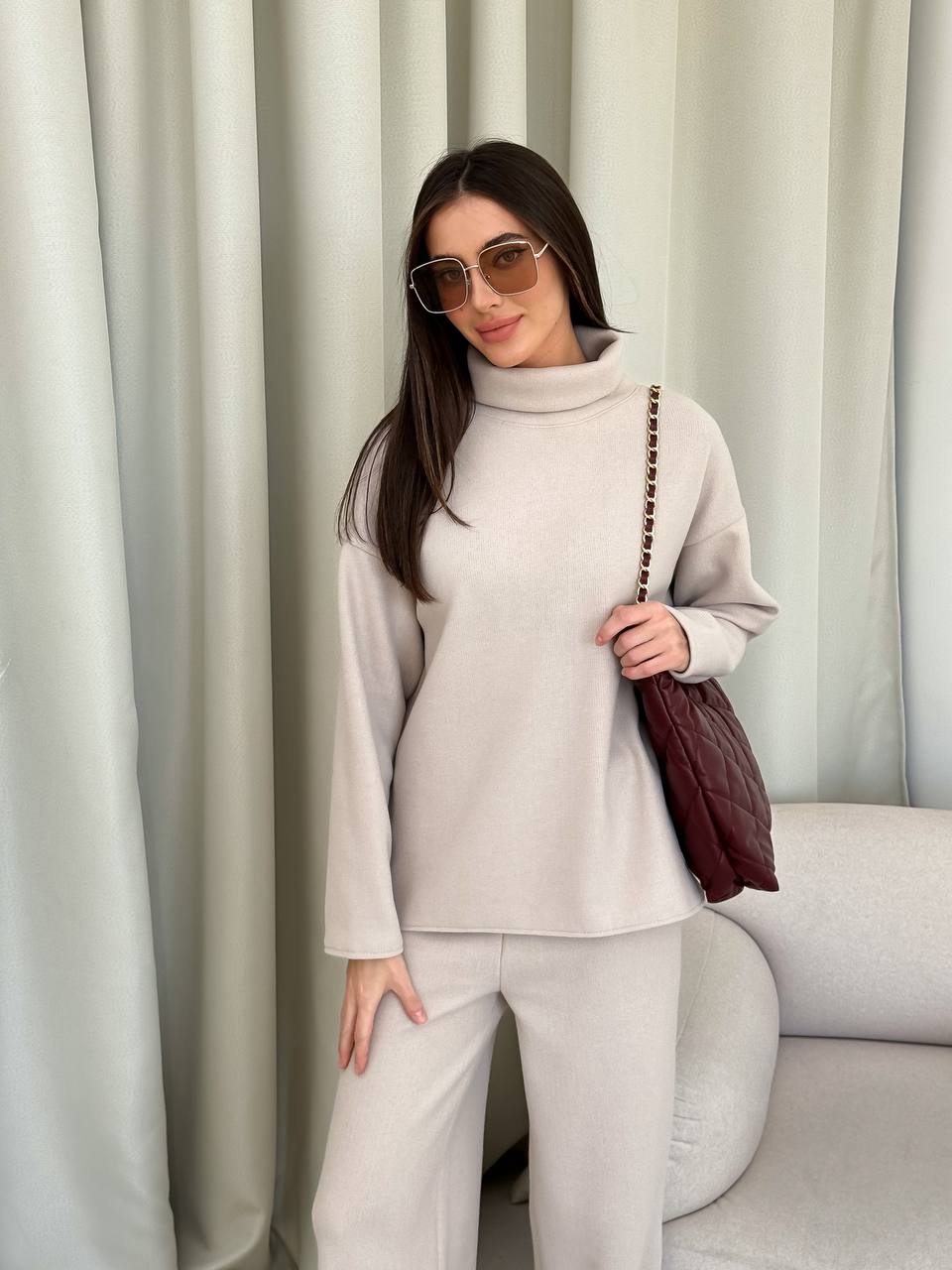 Woman in a light gray outfit holding a brown bag against a white curtain background
