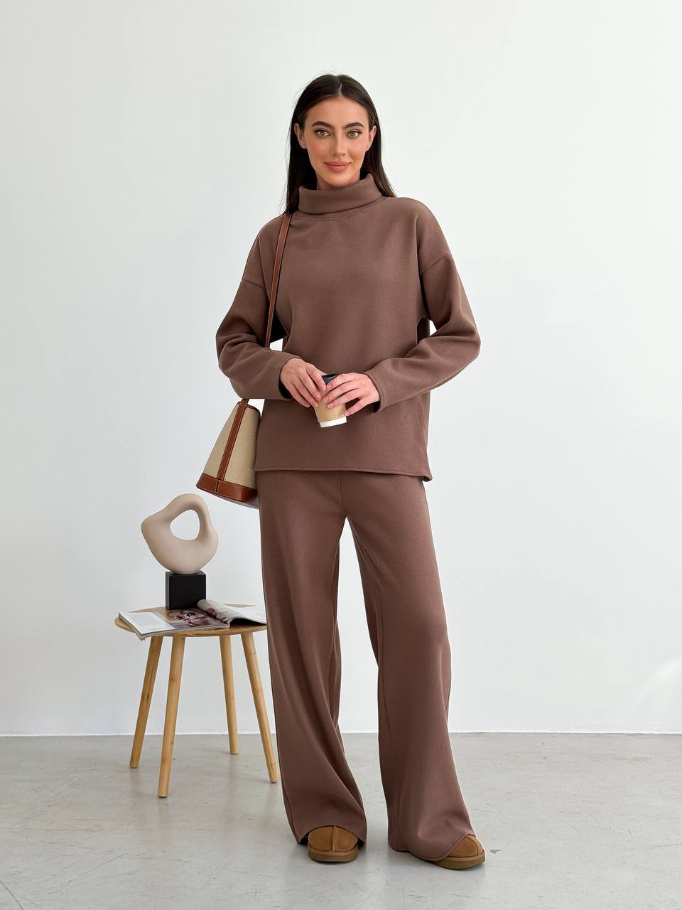 Woman wearing a brown outfit standing next to a chair with a bag on it against a white background