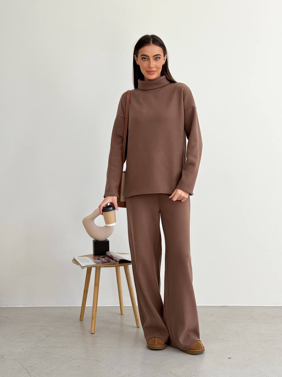 Woman in a brown outfit standing next to a stool with a coffee cup on a white background