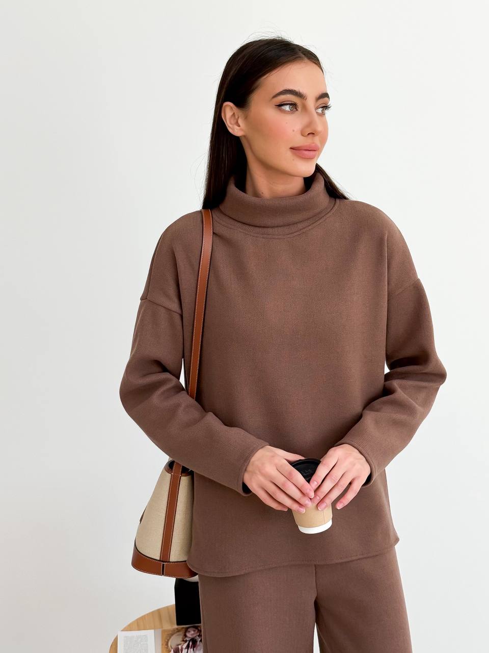 Woman wearing a brown turtleneck sweater holding a coffee cup against a white background