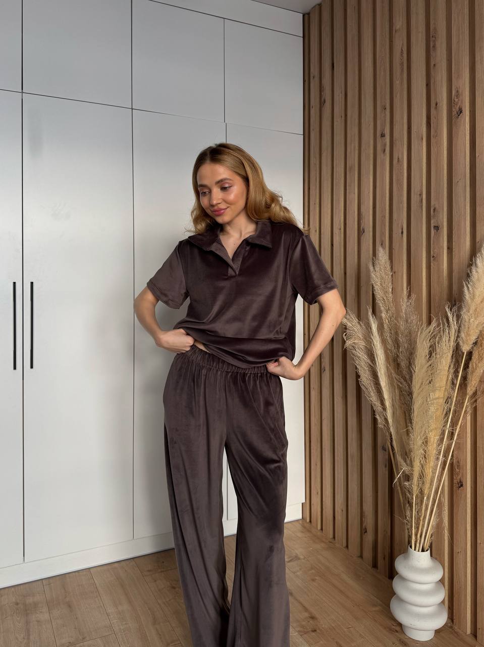 Woman in a brown outfit standing in a room with wooden walls and a vase of dried plants.